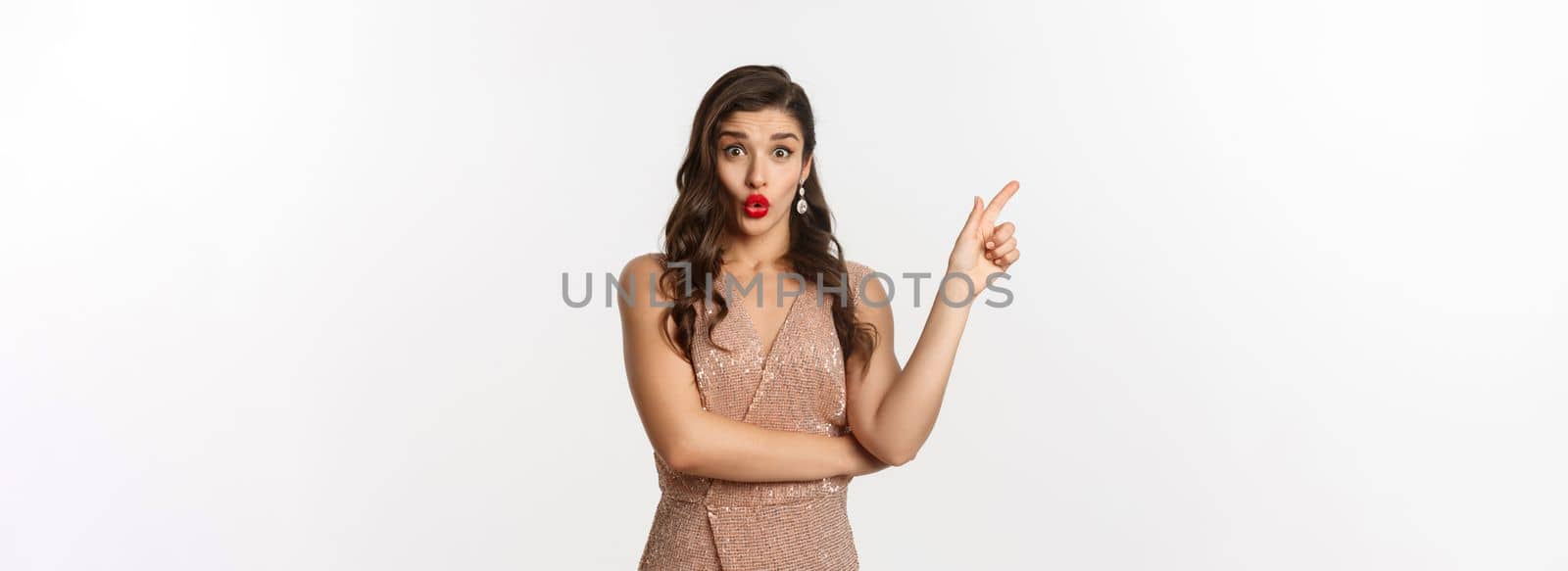 Christmas, holidays and celebration concept. Intrigued beautiful woman asking question about promo offer, pointing finger left at logo or banner, standing in party dress.