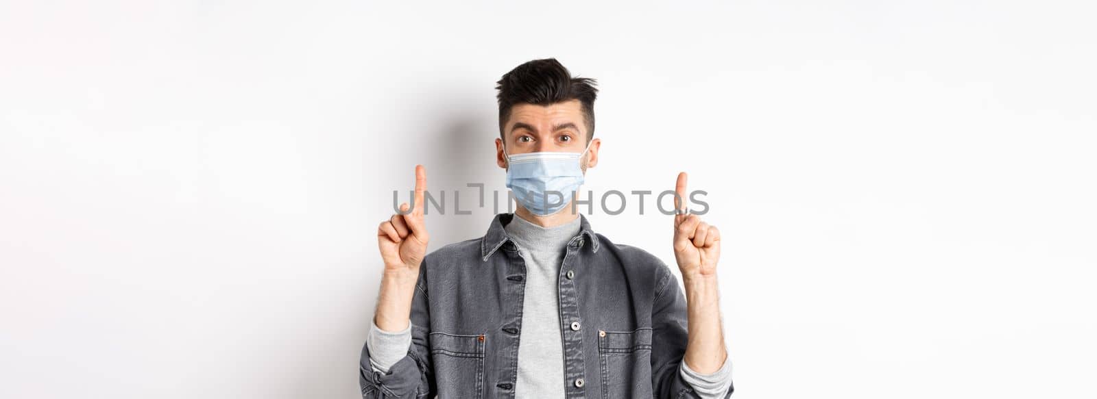 Pandemic lifestyle, healthcare and medicine concept. Stylish modern man in medical mask showing advertisement, pointing fingers up at logo, standing on white background by Benzoix