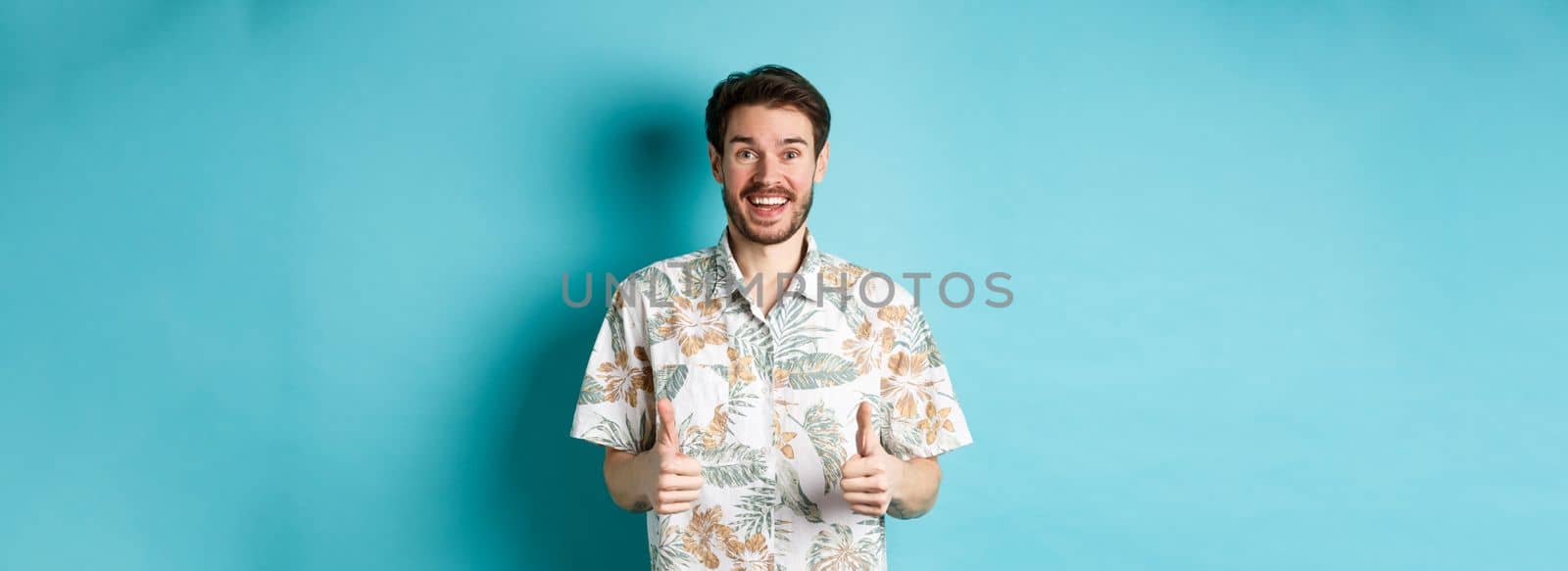 Summer holiday. Excited smiling tourist in hawaiian shirt show thumbs-up, praising hotel or vacation place, standing on blue background.