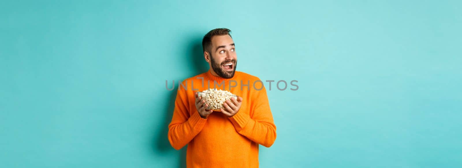 Excited and happy man watching tv and holding bowl of popcorn, looking left and smiling pleased, standing over blue background.