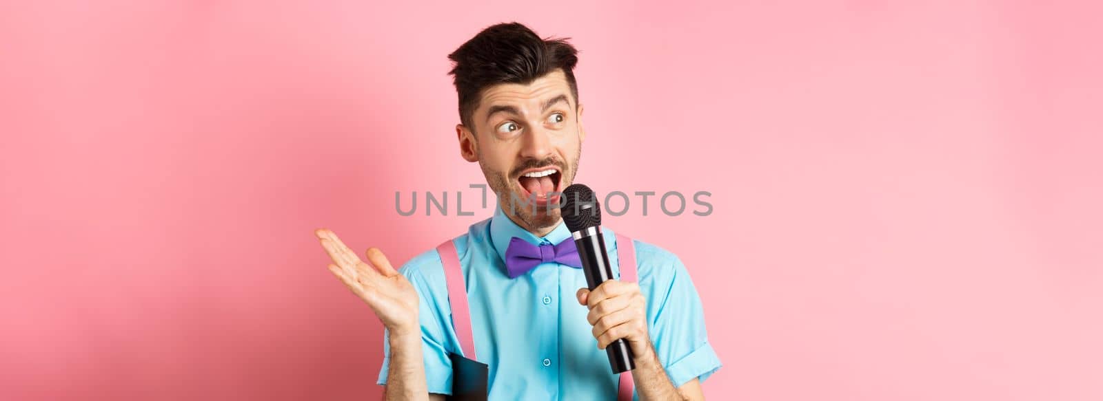 Image of male show host making speech, talking in microphone with clipboard under armpit, enteratin people on festive event, standing over pink background.