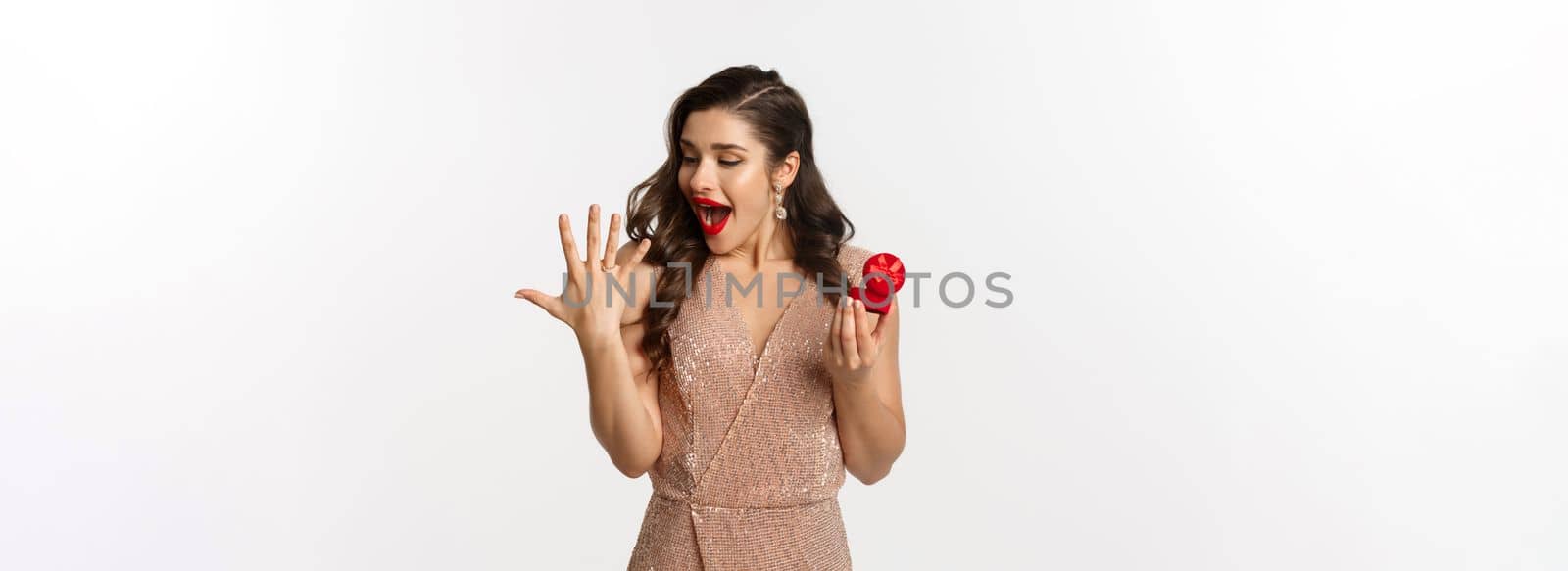Excited young woman in glamour dress being engaged, showing finger with ring and looking happy, receive marriage proposal, standing over white background.
