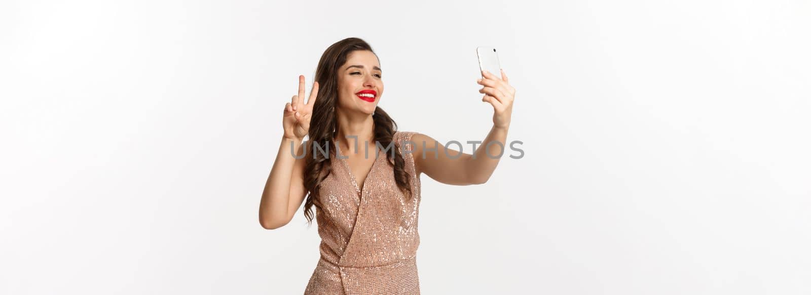 Attractive woman in luxury dress showing peace sign at phone camera, taking selfie on smartphone, posing in christmas party outfit, standing over white background.