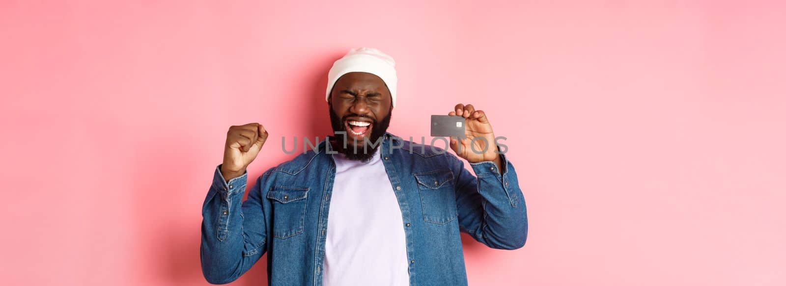 Shopping concept. Happy Black man rejoicing, scream of joy and showing credit card, standing over pink background.