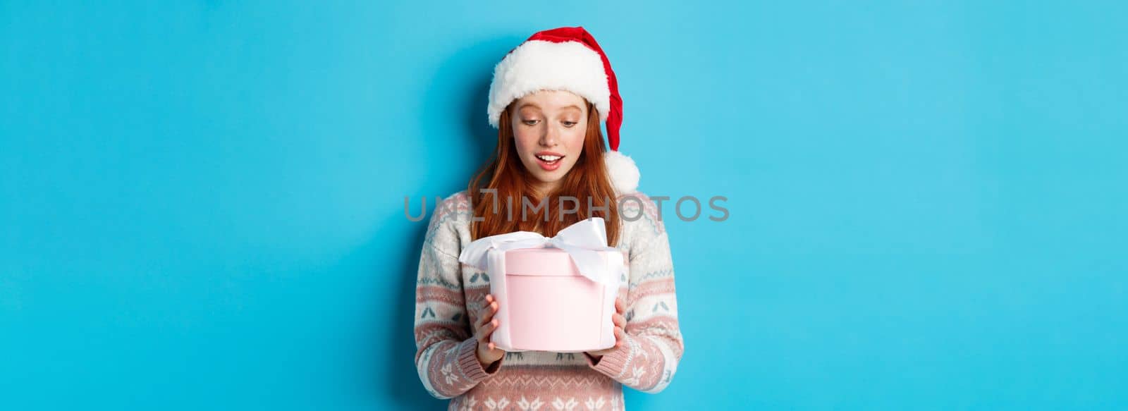 Winter and Christmas Eve concept. Touched and flattered redhead girl looking at box with xmas gift, smiling amazed, standing in santa hat against blue background by Benzoix