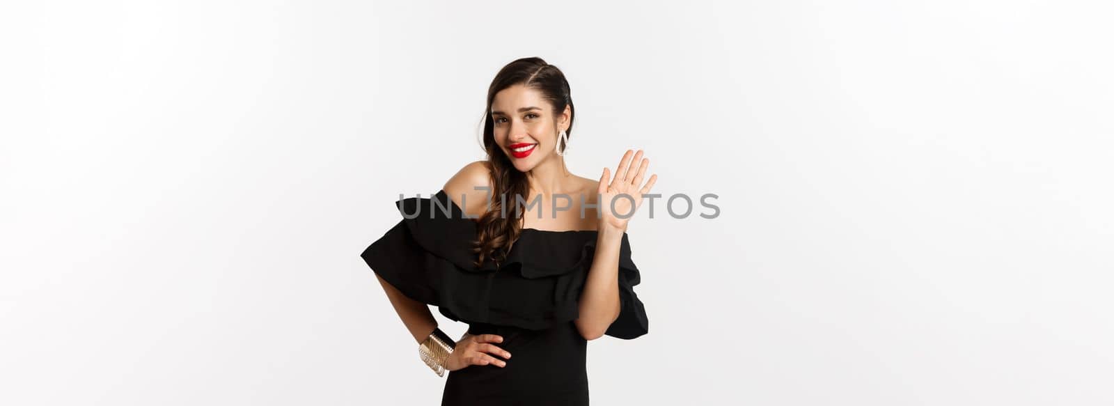 Gorgeous young woman in black dress, earrings and jewelry, waving hand and smiling friendly, saying hello, standing over white background.