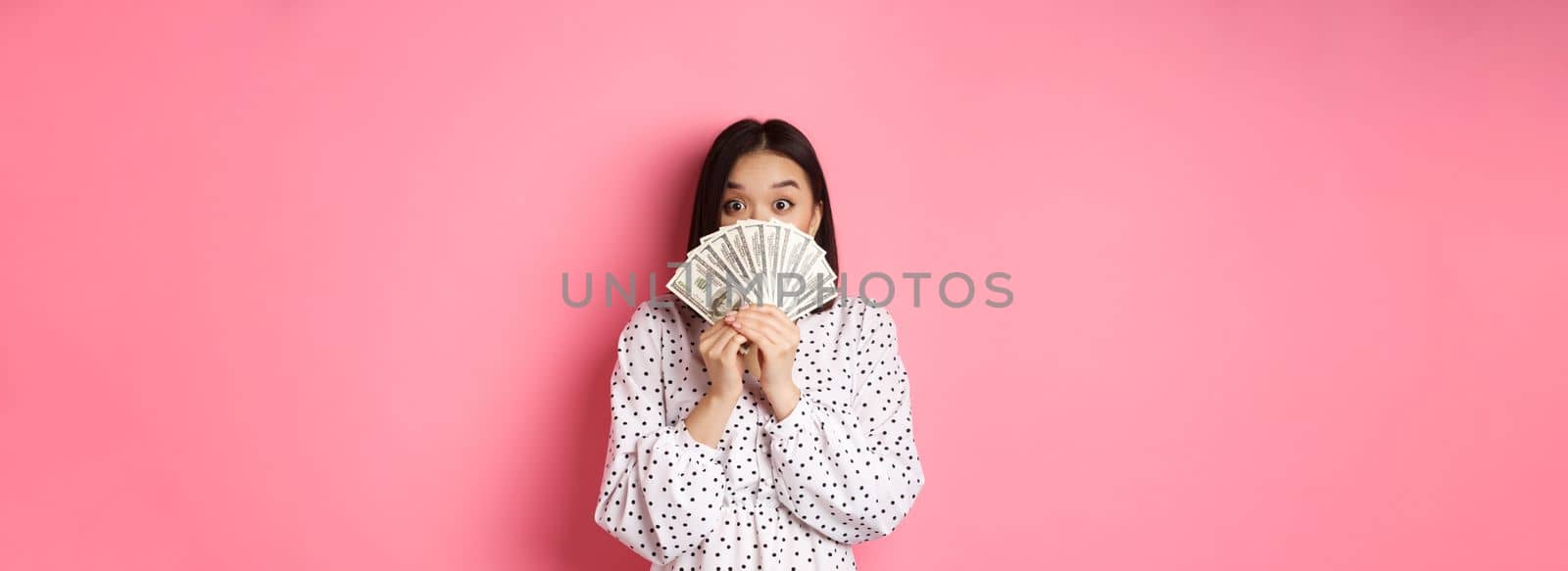 Shopping concept. Cute asian woman hiding face behind money dollars, peeking at camera, standing over pink background.