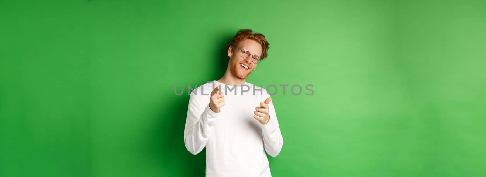 Cheeky young man in glasses praising and congratulating you, pointing fingers at camera and smiling, standing in glasses and white long-sleeve against green background.