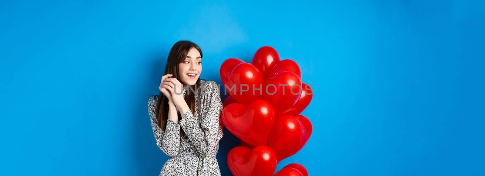Valentines day. Beautiful romantic girl dreaming off date, standing near lovely heart balloons and smiling, blue background.