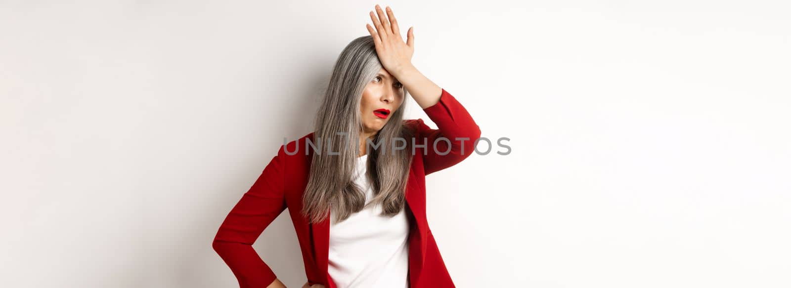 Annoyed asian office lady in red blazer roll eyes and facepalm, standing bothered and irritated over white background.