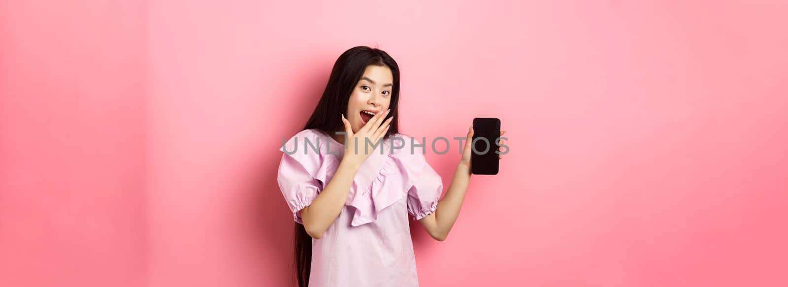 Cheerful asian teen girl showing empty phone screen, laughing and covering mouth with hand, standing in dress against pink background by Benzoix