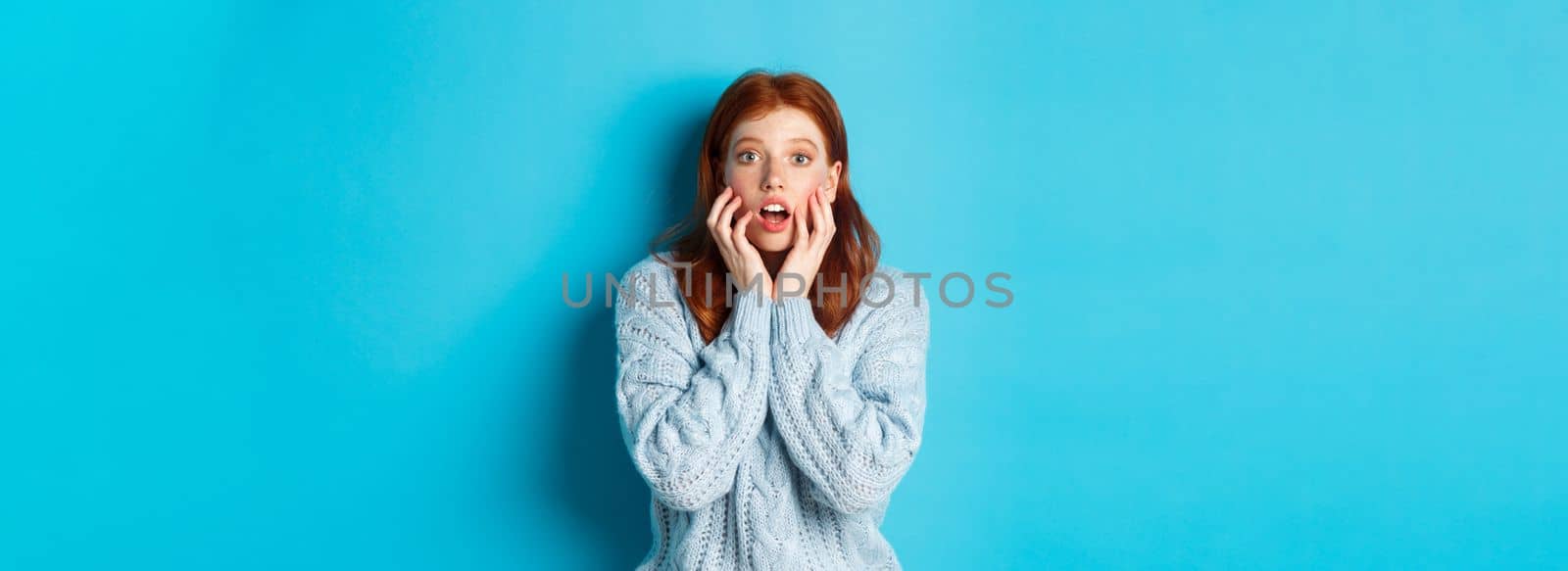 Startled redhead girl staring at something amazing, gasping in awe, standing in sweater against blue background. Copy space