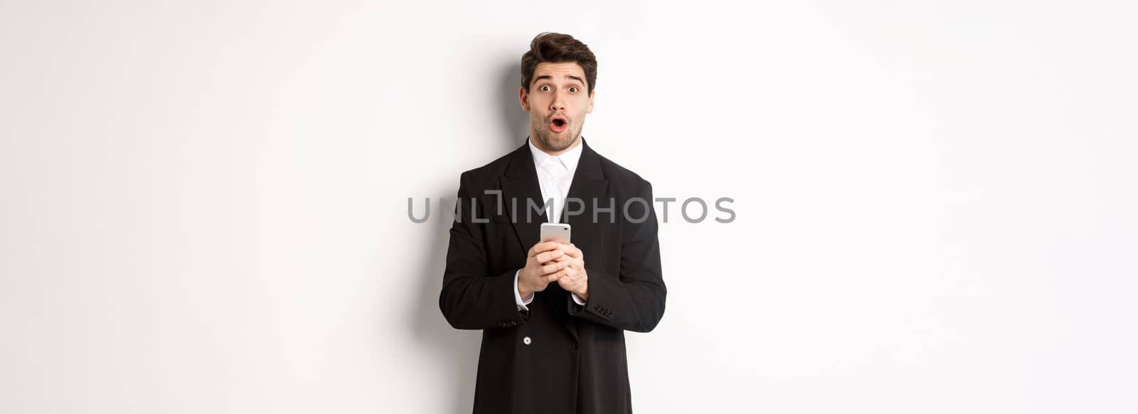 Amazed handsome guy in black suit reacting to cool promo offer, holding mobile phone, standing against white background.