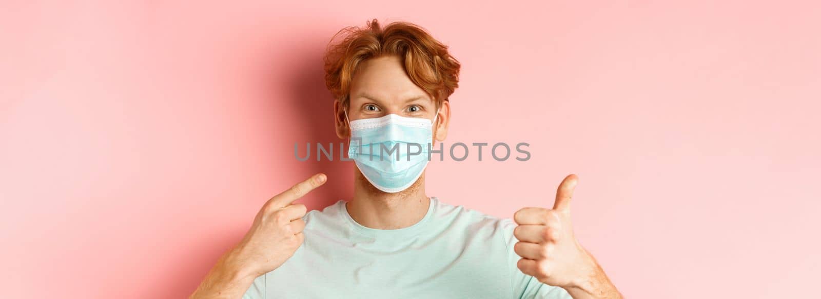 Covid-19 and pandemic concept. Handsome redhead guy pointing finger at face mask and showing thumbs-up, using measures from coronavirus, standing over pink background.