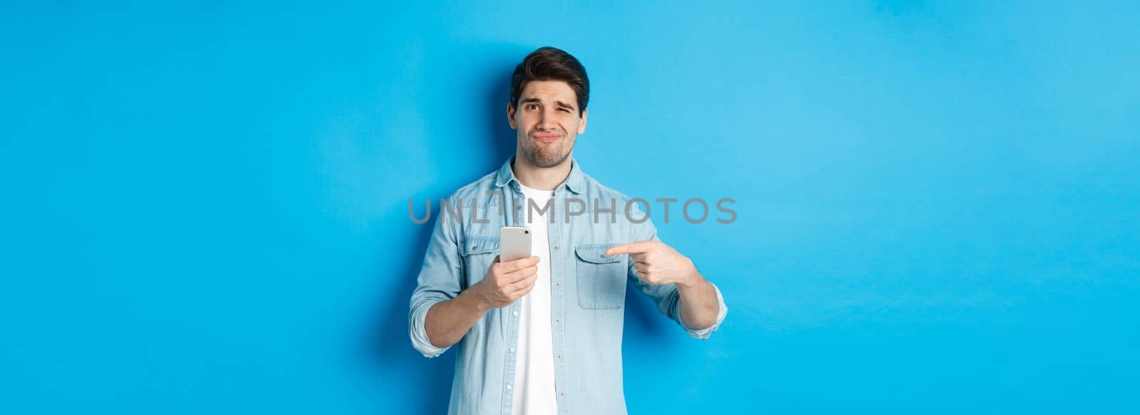 Concept of online shopping, applications and technology. Skeptical and displeased guy pointing finger at smartphone and grimacing disappointed, standing over blue background by Benzoix