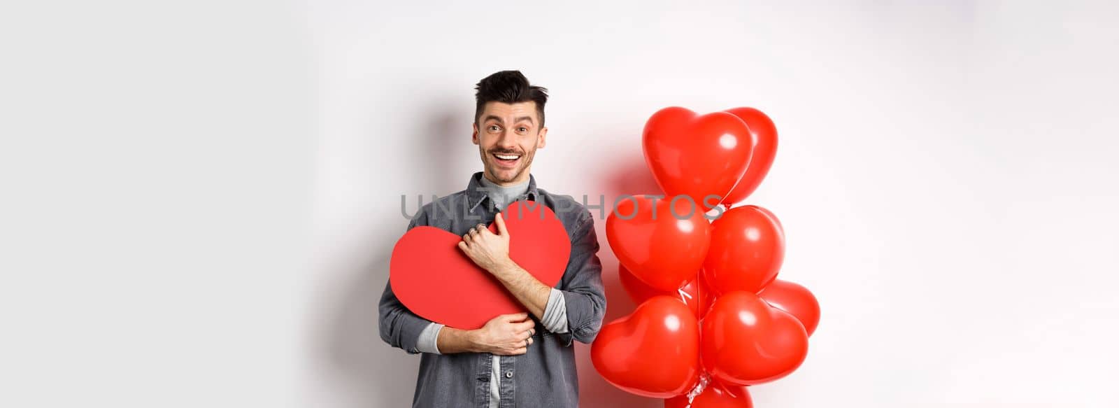 Young romantic man looking with love at camera, hugging Valentine heart card and smiling happy, celebrating lovers holiday, standing near red balloons, white background.
