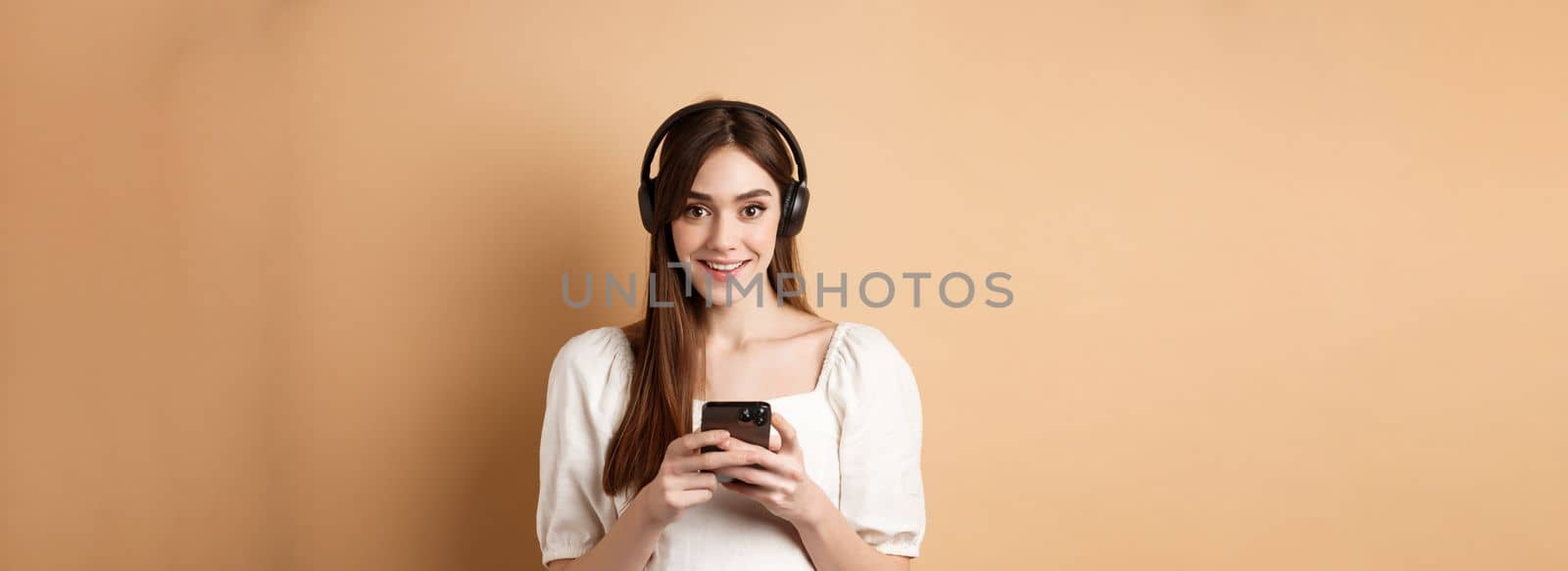 Pretty girl in headphones smiling at camera, listening music and using mobile phone app, beige background.
