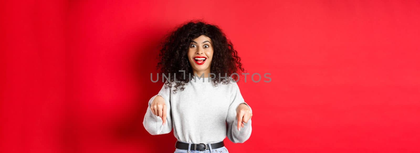 Excited woman pointing fingers down and smiling amazed, look here gesture, showing logo on red empty space by Benzoix