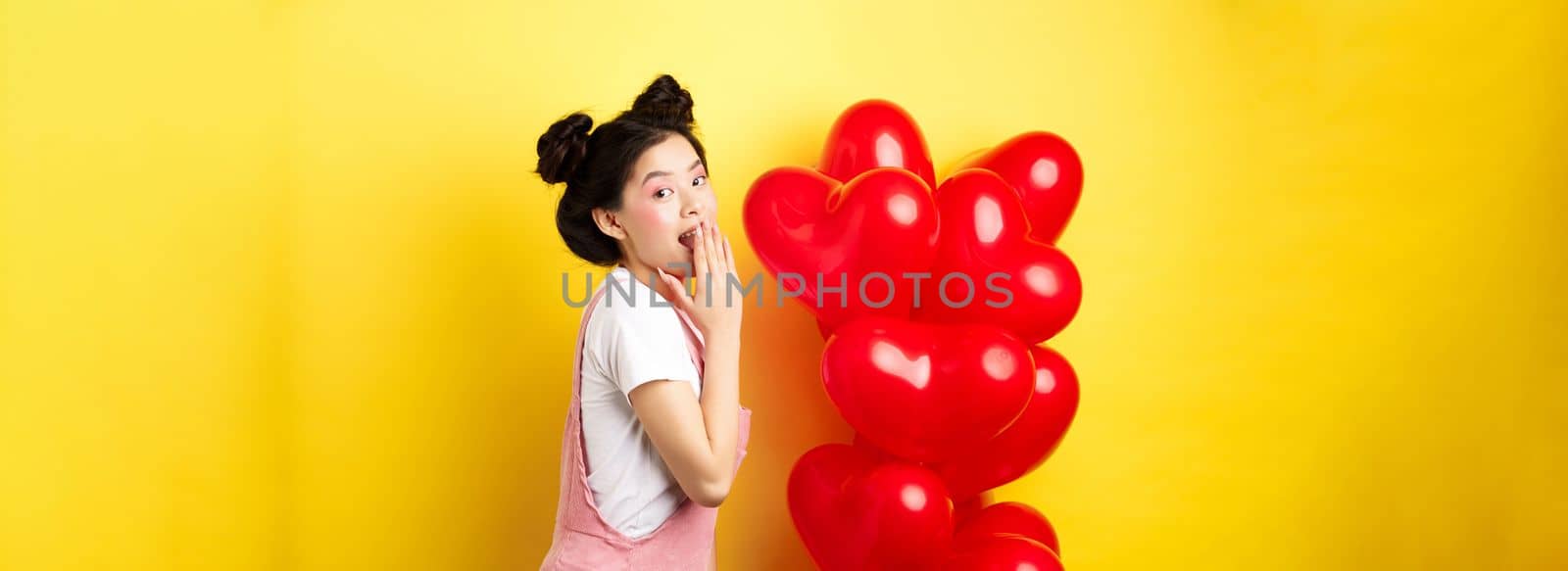 Valentines day and relationship concept. Coquettish and romantic girl laughing, covering mouth with hand, look silly at camera, standing near red heart balloons, yellow background.