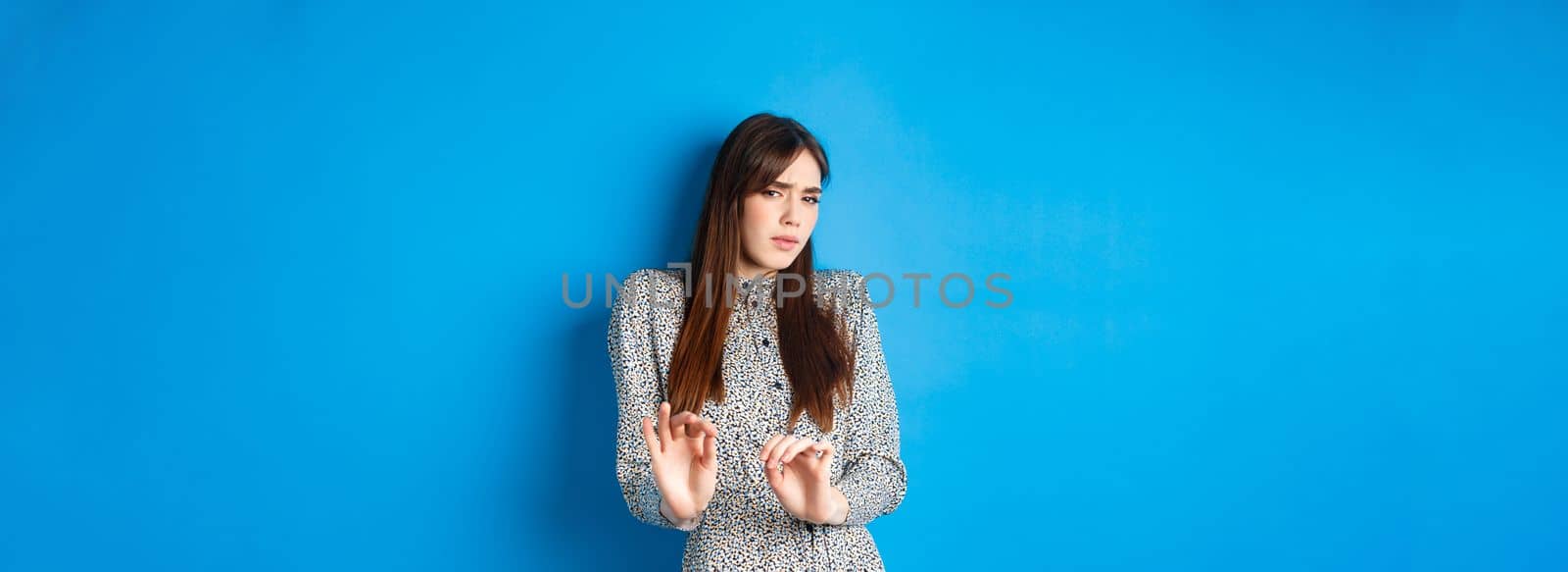 Disgusted and reluctant woman asking to keep away from here, showing block gesture, raising hands to stop you, standing on blue background.