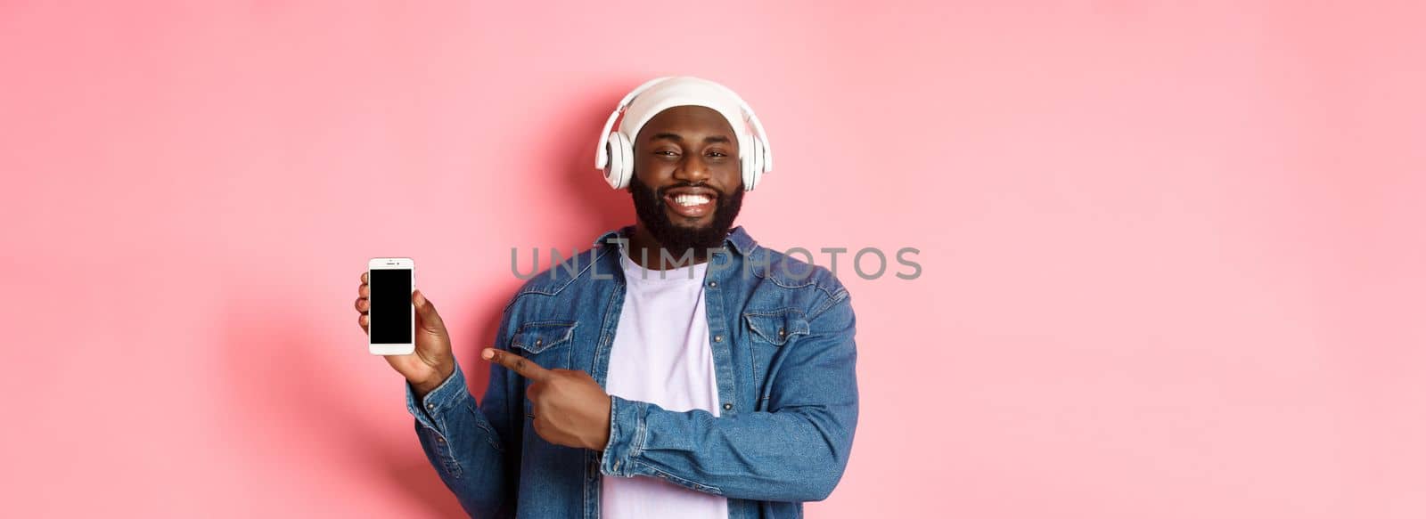 Happy black man listening music in headphones and smiling, pointing mobile phone screen app or playlist, standing over pink background.