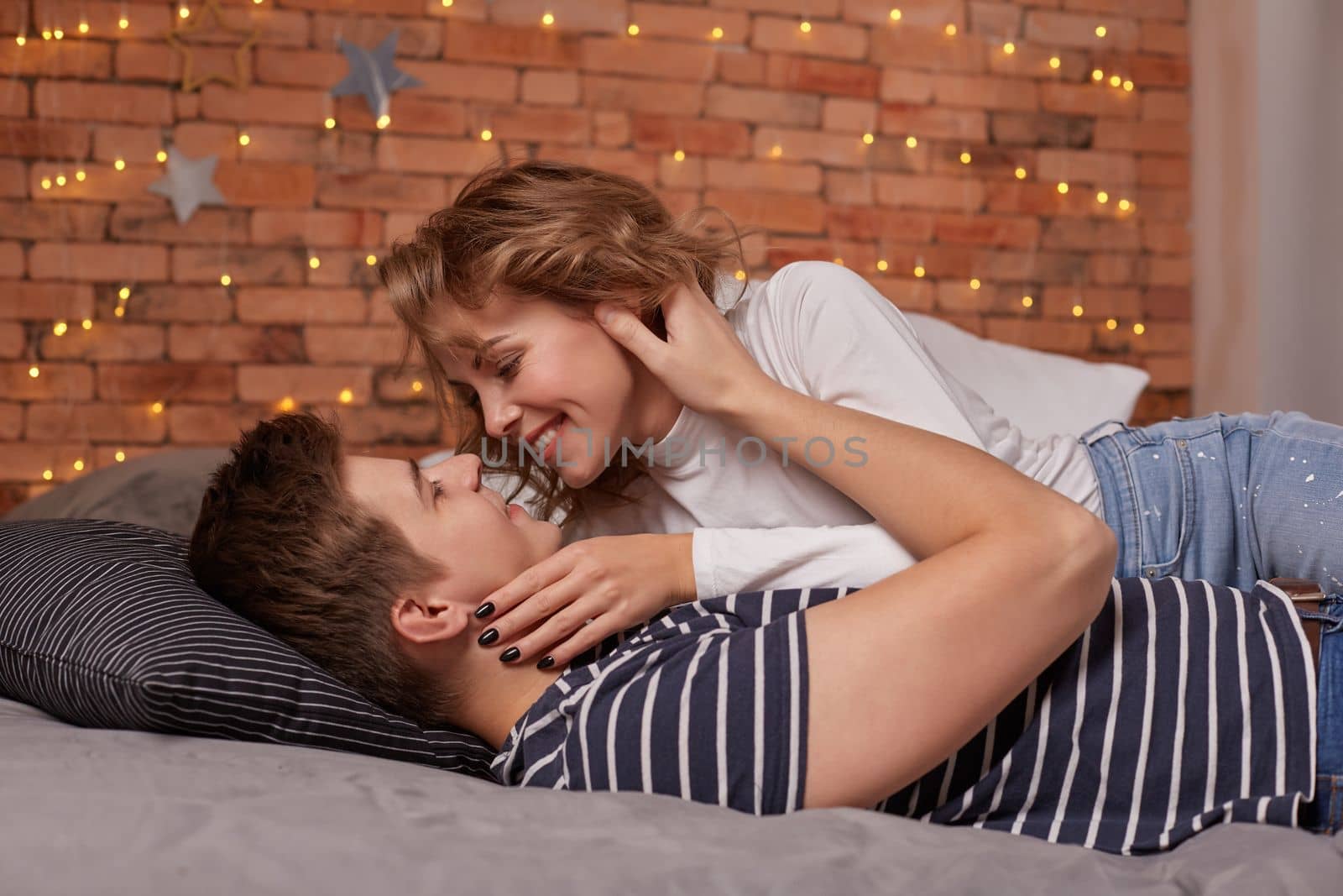 Caucasian young beautiful couple lie on bed and hugging each other. They look each other in the eyes and they are happy