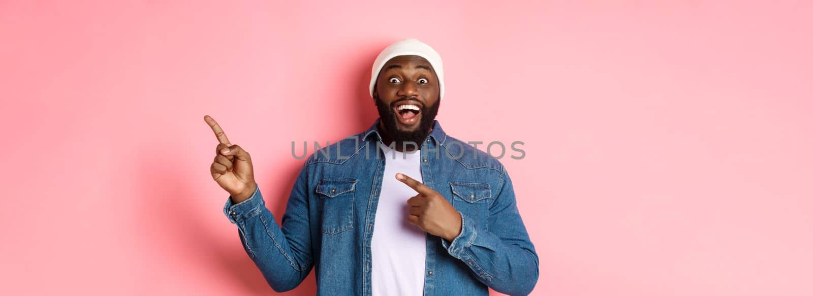 Excited and amazed Black man showing awesome offer, pointing fingers right at copy space, standing in hipster beanie and denim shirt on pink background.