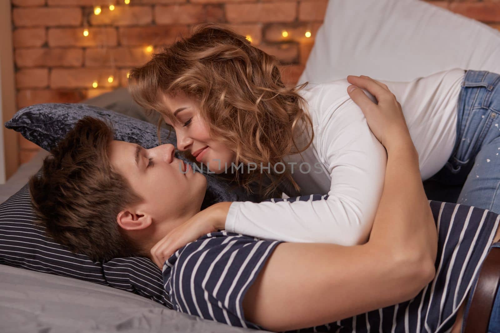 Caucasian young beautiful couple lie on bed and hugging each other. They look each other in the eyes and they are happy