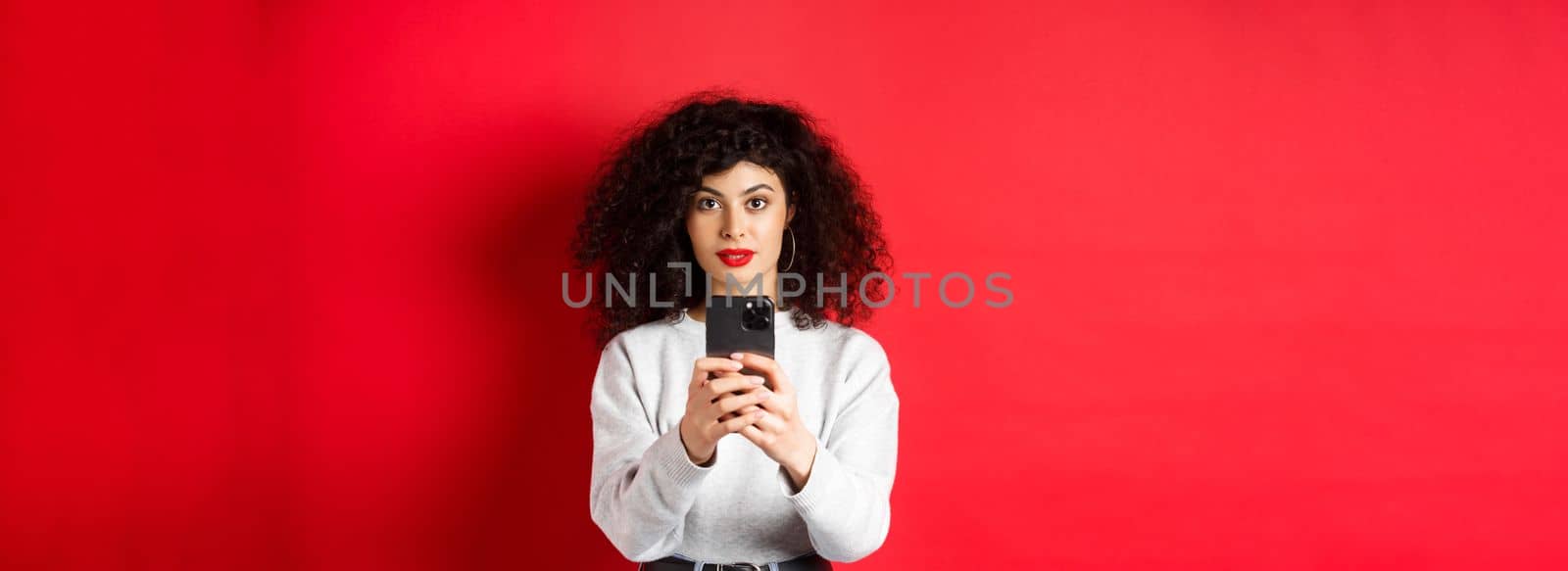 Young woman with curly hair, recording video on smartphone, taking photo on mobile phone and looking at camera, standing on red background by Benzoix