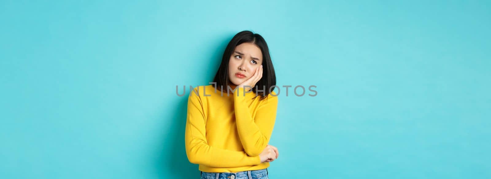 Sad and bored asian teenage girl leaning on palm, looking at upper left corner with lonely and upset expression, standing over blue background.