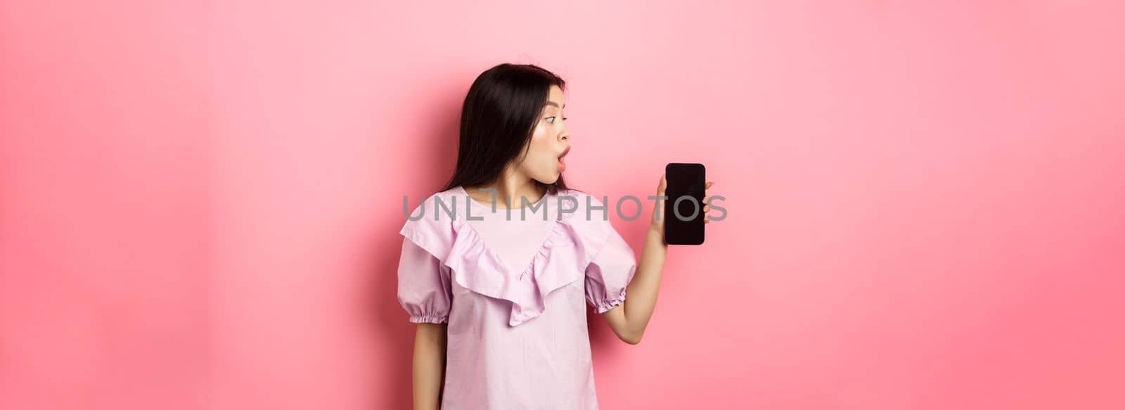 Wow look here. Excited asian girl look at smartphone screen with amazed face, checking out online offer, standing against pink background by Benzoix