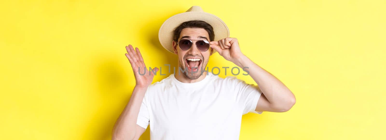 Concept of tourism and holidays. Close-up of surprised man shouting for joy, enjoying vacation, wearing sunglasses with summer hat, yellow background by Benzoix