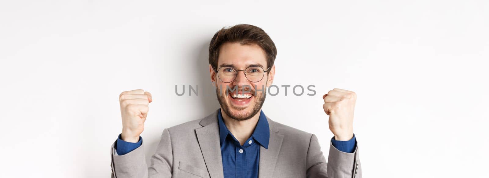 Close-up of cheerful male ceo manager in glasses and suit, saying yes and celebrating win, making fist pump, standing on white background by Benzoix
