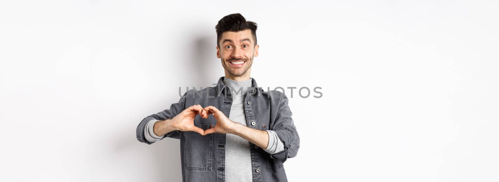 Smiling boyfriend showing heart with love, looking romantic and happy at lover, standing on white background. Valentines day and relationship concept.