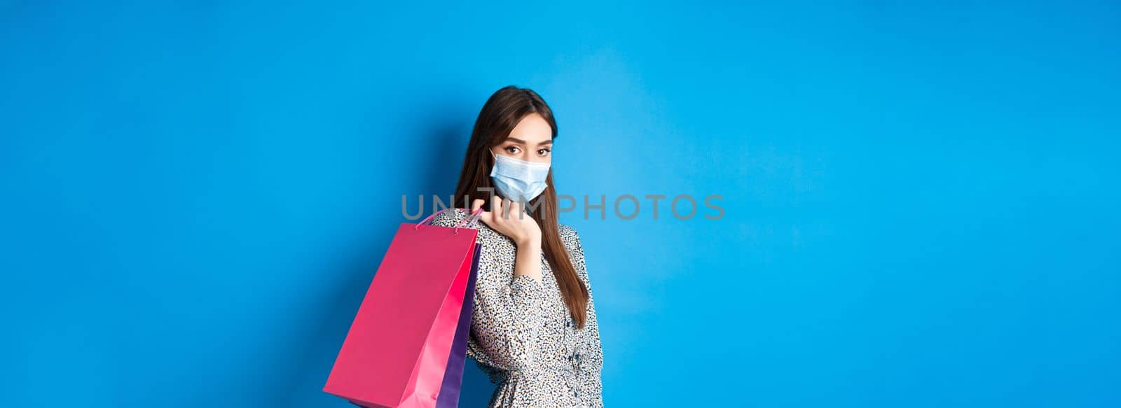 Covid-19, pandemic and lifestyle concept. Elegant woman wear medical mask on shopping, holding bags over shoulder and walking against blue background.