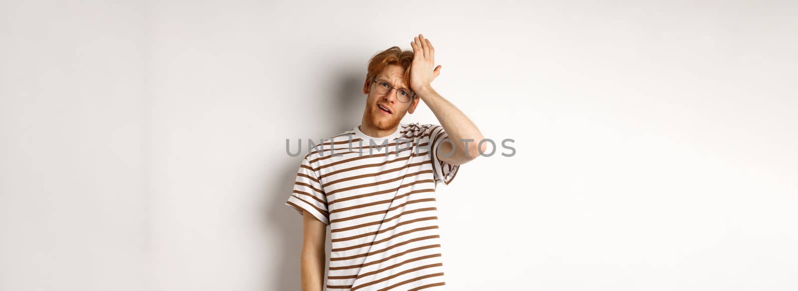 Tired and distressed redhead male student slap forehead, making facepalm and staring at camera exhausted, standing over white background.