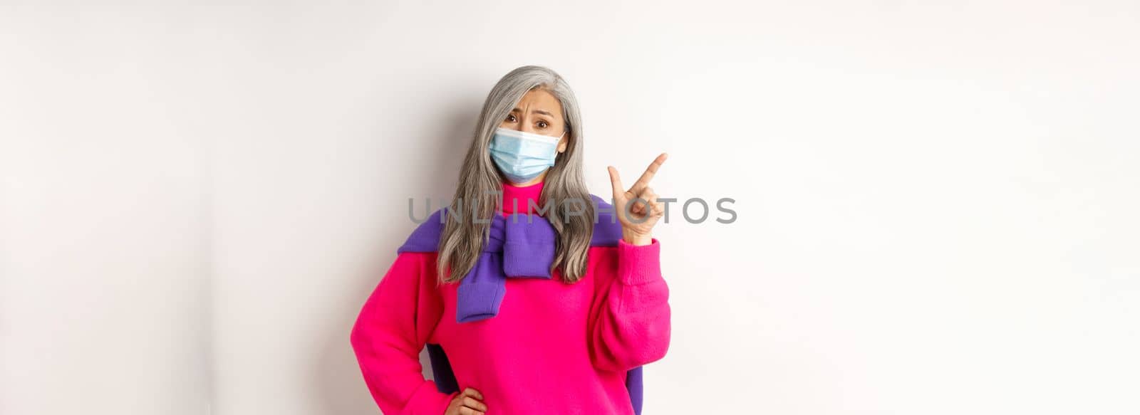 Covid-19, quarantine and lockdown concept. Skeptical asian elderly female in face mask looking disappointed, pointing at upper left corner, white background.