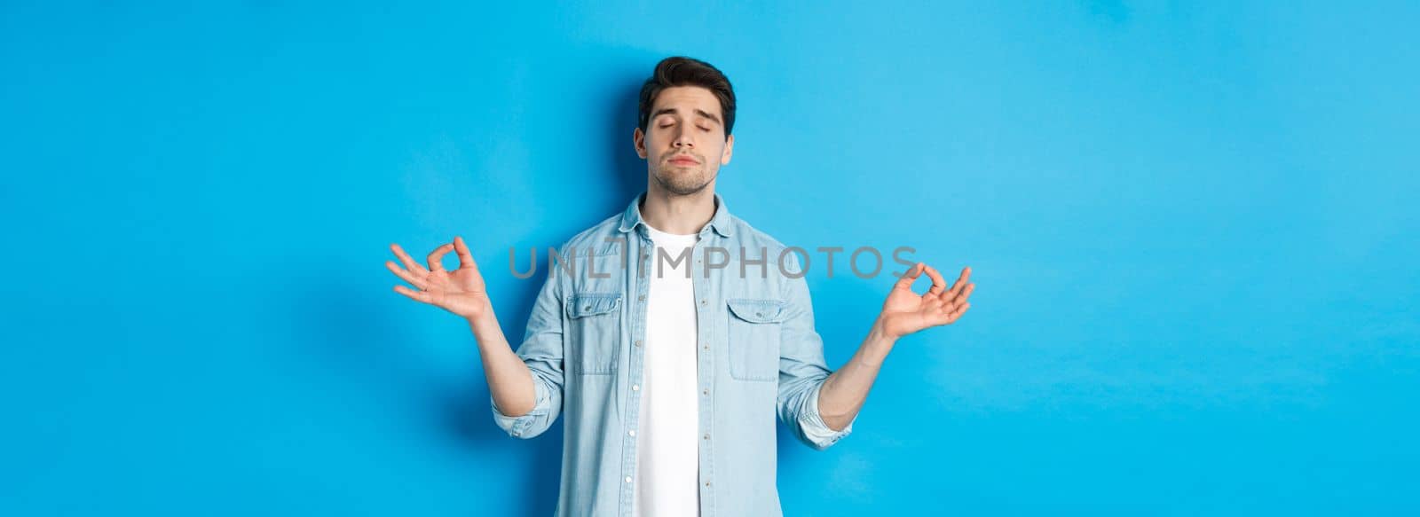 Calm man with closed eyes meditating, holding hands sideways and do yoga breathing exercises, standing against blue background.