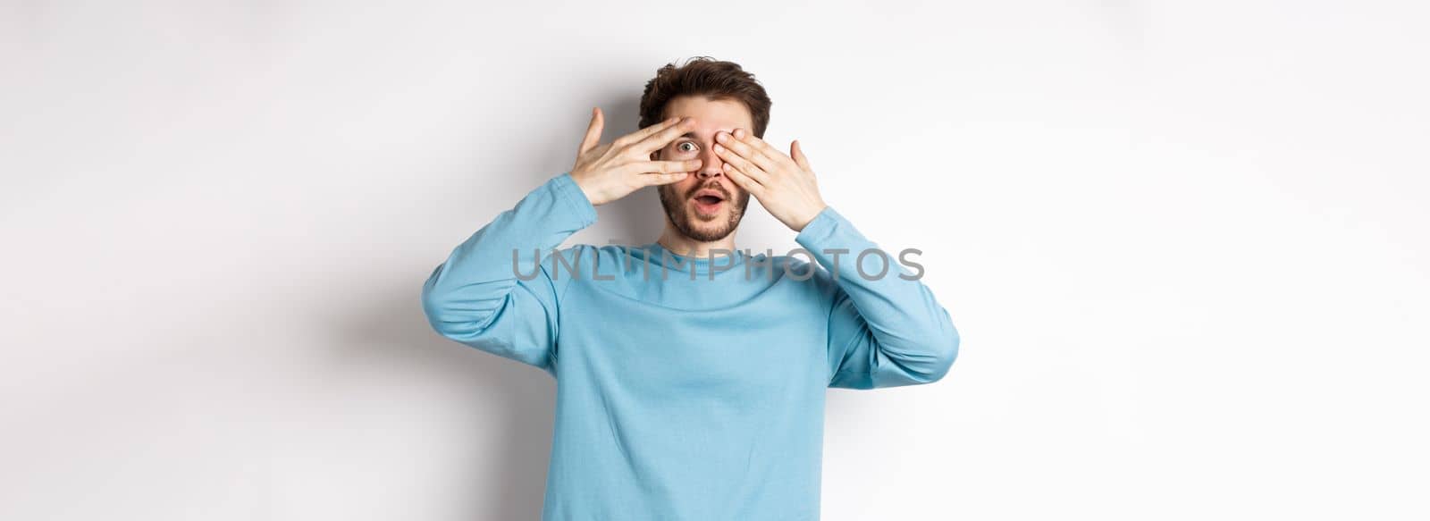 Amazed smiling man covering eyes with hands but peeking through fingers at something awesome, standing over white background.