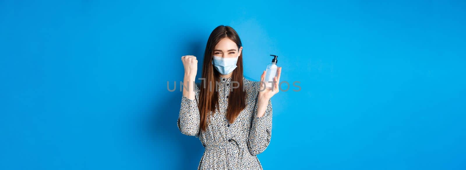 Covid-19, social distancing and healthcare concept. Motivated and excited girl in medical mask cheering, showing bottle of hand sanitizer and say yes, standing on blue background.