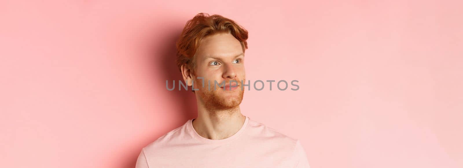 Handsome european male model with red hair and beard, turn head and looking pleased at copy space on left side, standing over pink background.