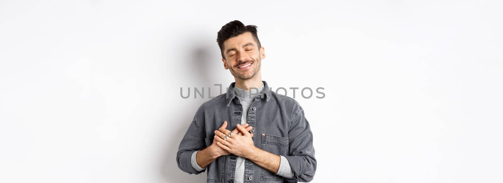 Romantic happy man holding hands on heart and smiling with closed eyes, dreaming of something, feeling nostalgic from sweet memories, standing on white background.