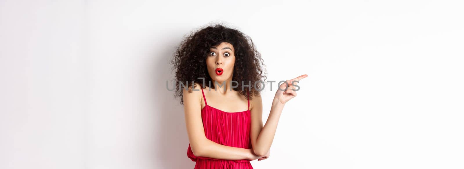 Surprised elegant woman in red dress, saying wow and pointing finger left, showing interesting promo, standing over white background by Benzoix