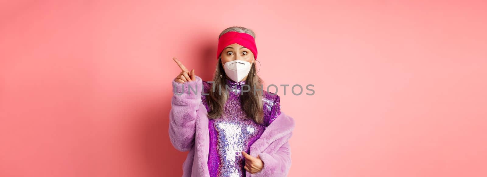 Coronavirus and shopping concept. Amazed asian senior woman in face mask staring at camera, pointing upper right corner promo, standing over pink background by Benzoix