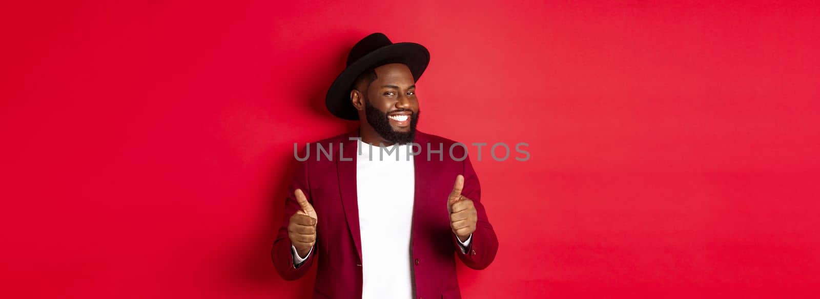 Cheerful Black man having fun on party, showing thumbs up in approval, smiling and liking something, standing against red background by Benzoix