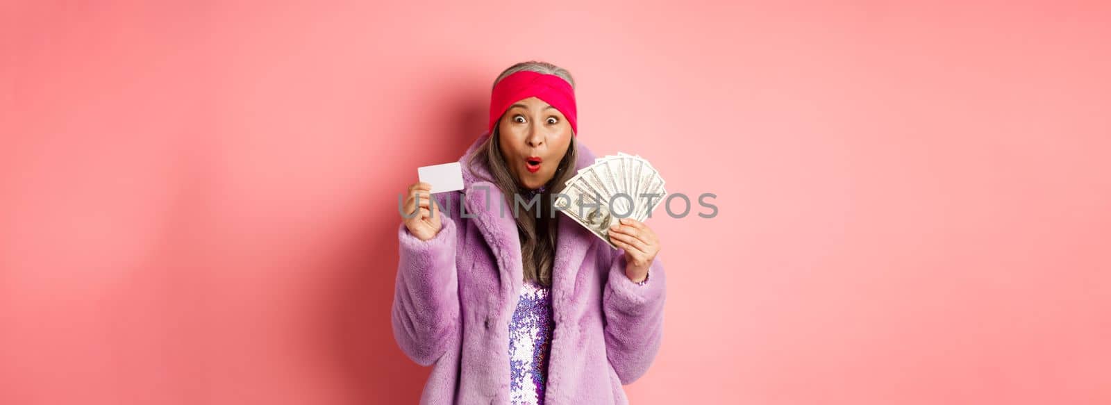 Shopping and fashion concept. Happy senior asian woman holding money in dollars and plastic card, looking amazed and ecstatic, pink background by Benzoix