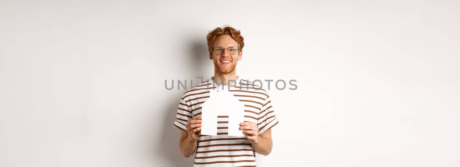 Handsome young man with beard and red hair, wearing glasses and striped t-shirt, showing paper house cutout and smiling, concept of real estate and buying property.