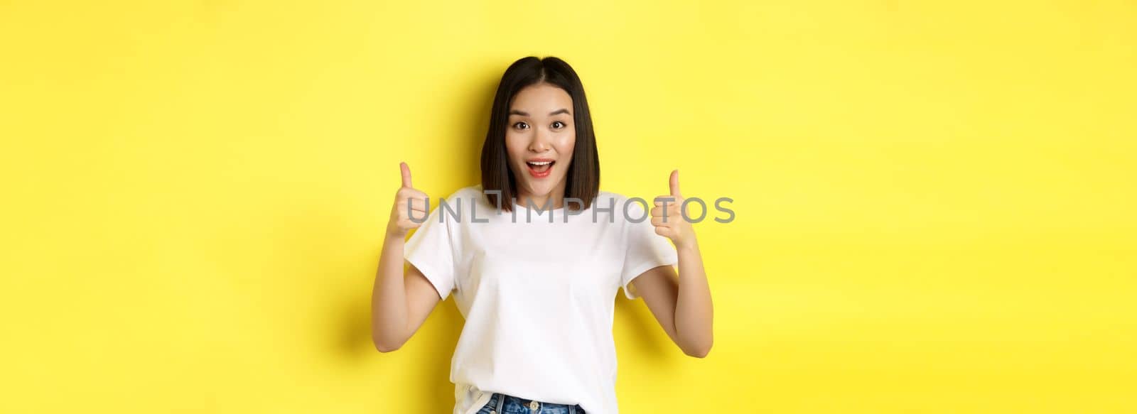 Pretty young asian woman in white t-shirt, showing thumbs up and smiling, praise good offer, recommend product, standing satisfied over yellow background by Benzoix