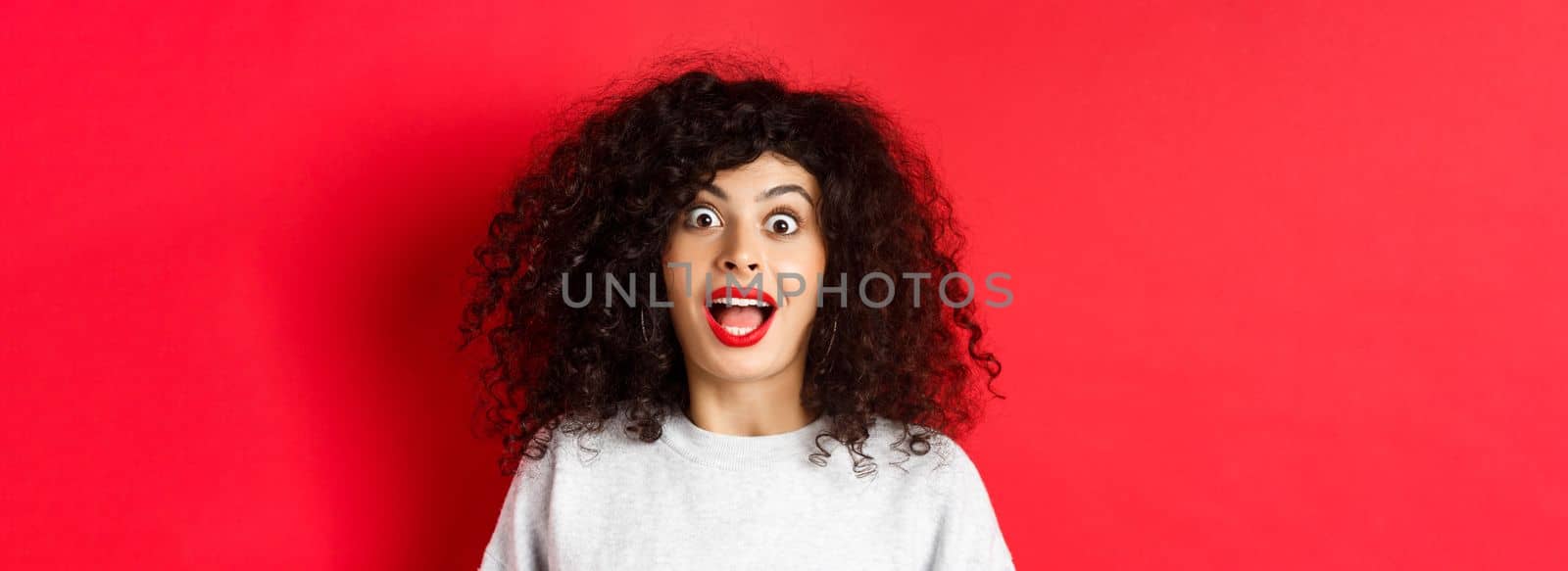 Close-up portrait of excited woman with curly hair, scream surprised and amazed, checking out special deal, standing on red background.
