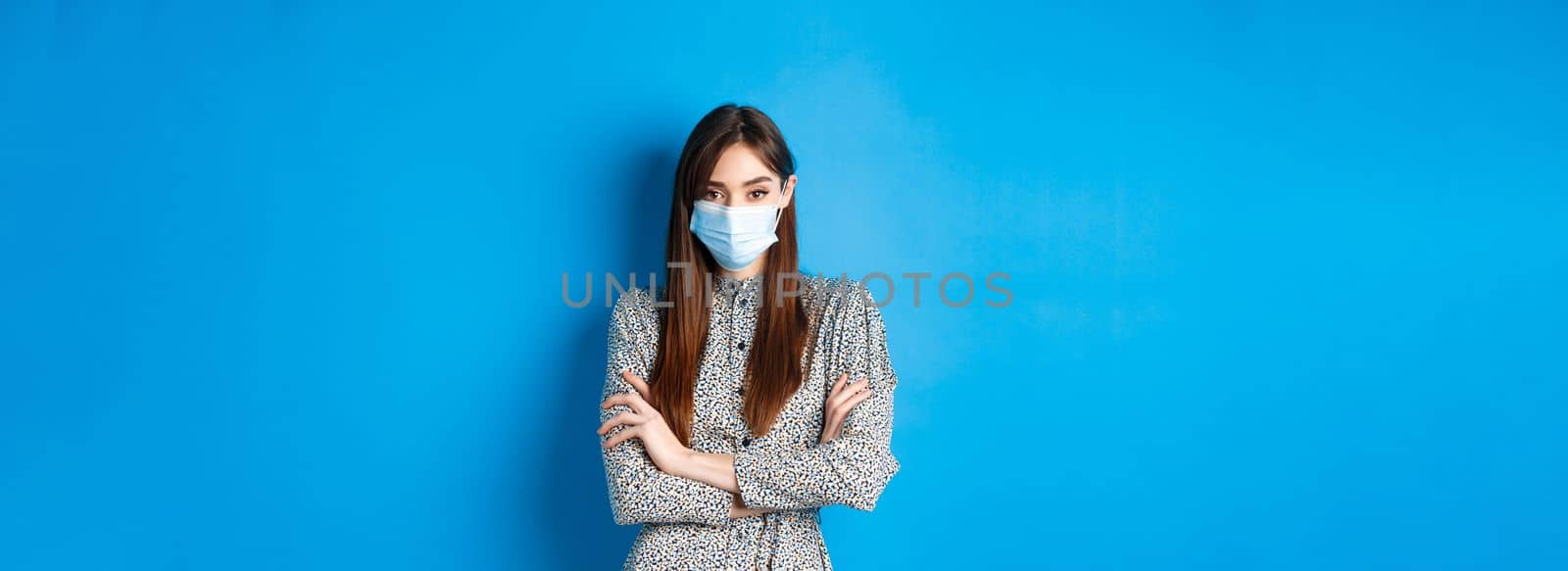 Covid-19, pandemic lifestyle concept. Serious young woman with long natural hair, wearing medical mask, cross arms on chest, protect herself from virus, blue background.
